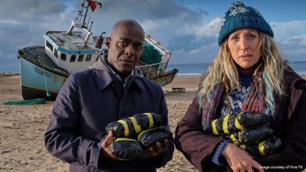 Paterson Joseph as Samuel, and Daisy Haggard as Janet, standing on beach in front of grounded boat in still from BBC’s Boat Story